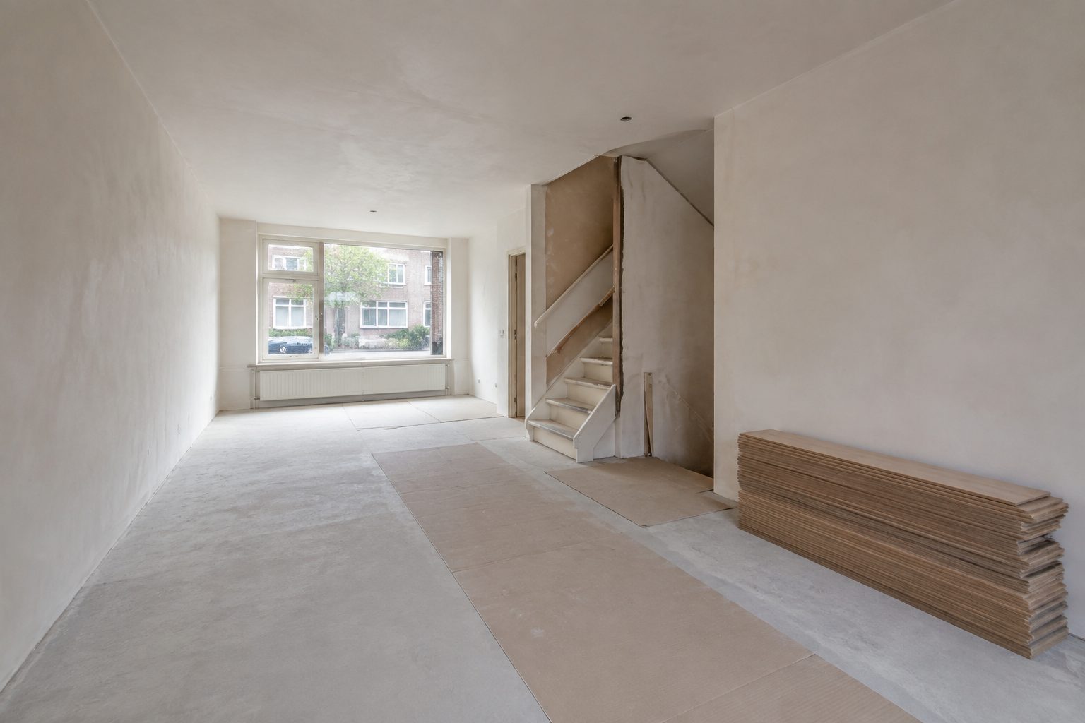 Woonkamer in renovatie met gestucte wanden, kale dekvloer en houten planken die acclimatiseren